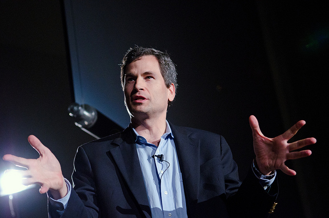 A person wearing a suit speaks animatedly, making hand gestures, in front of a microphone