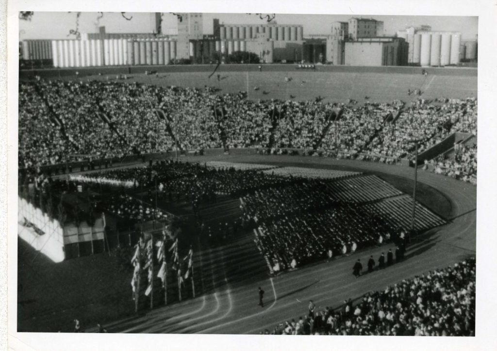 Graduates sit in chairs on the field of Memorial Stadium in front of a stage. Attendees are in the stadium bleachers.