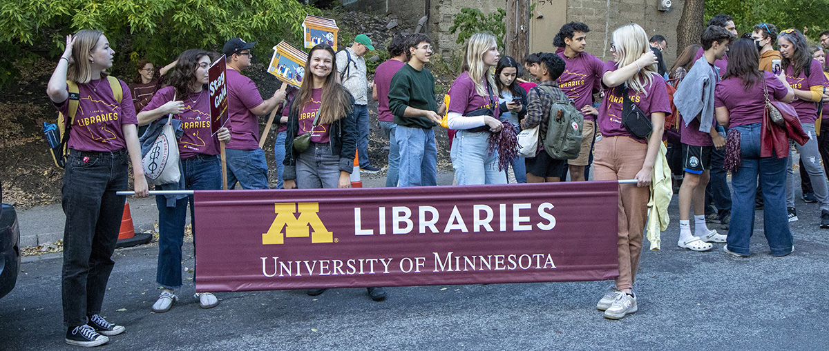 Several students wearing matching maroon University of Minnesota Libraries t-shirts gathered on the street in front of a banner that reads "University of Minnesota Libraries" 