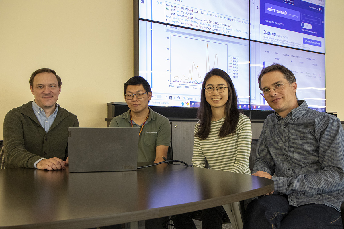 Four researchers sit side by side at a table; in front of them is an open laptop, and behind them a screen displaying data