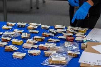 Dozens of brightly colored boxes that contain pharmaceutical products are on a table covered by a blue-quilted cloth. Anna Shepherd's blue-gloved hand is seen pointing at a box on the table.
