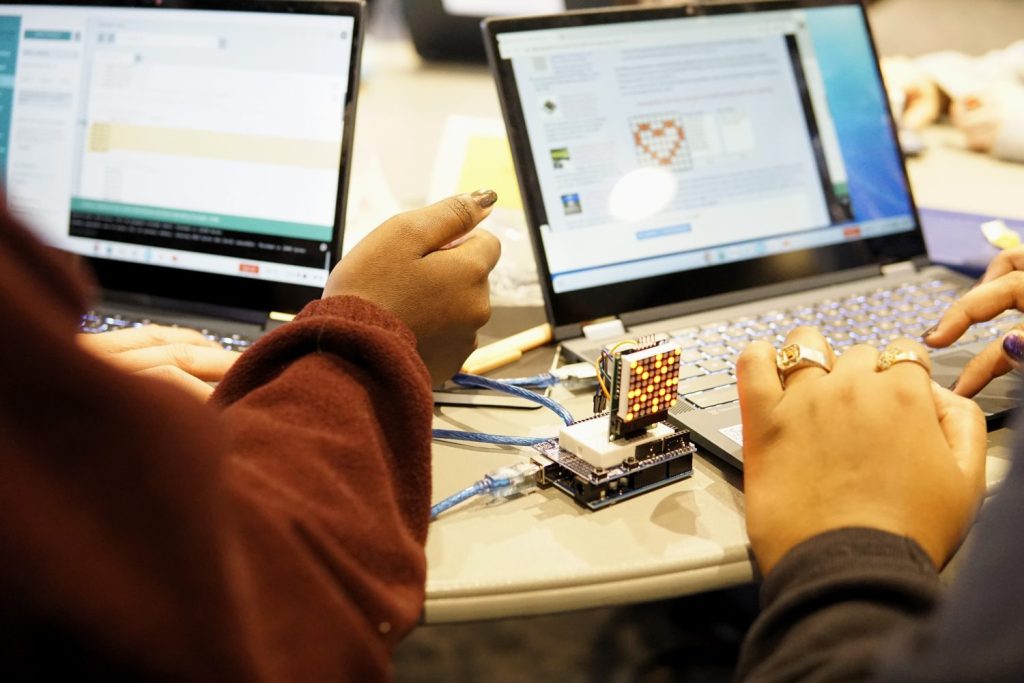 Two students work on laptops. One is looking at an Arduino code page, and the other is designing an LED matrix pattern in a heart shape. An LED matrix showing an asterisk pattern is plugged in to one of the laptops.