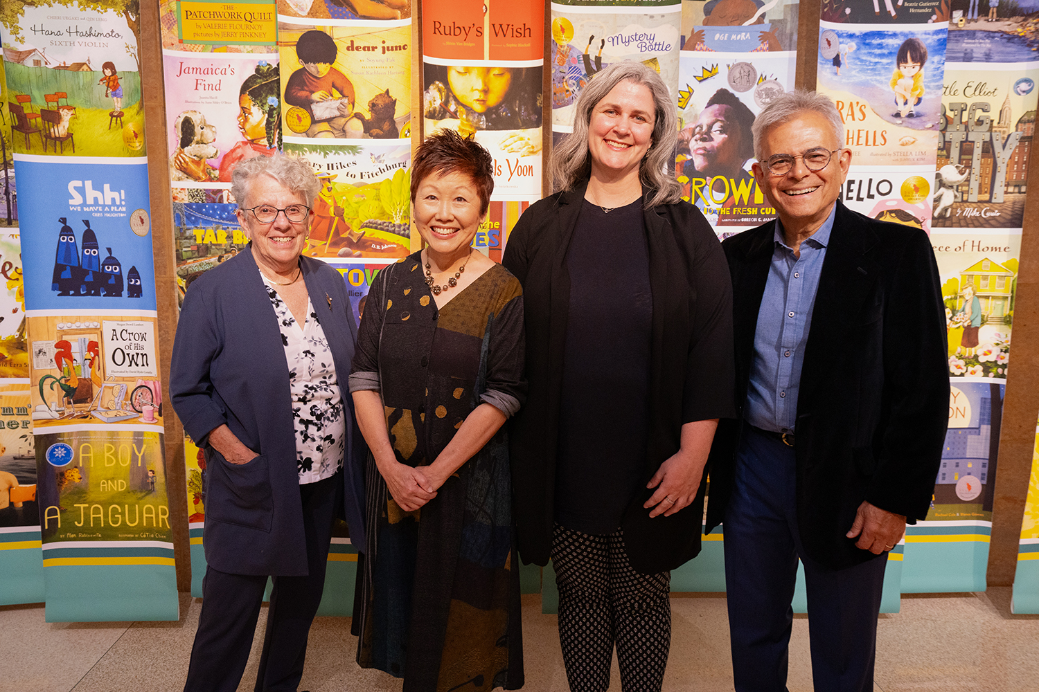 Four people stand side by side in front of a wall covered in images of colorful children's book covers at Elmer L. Andersen Library