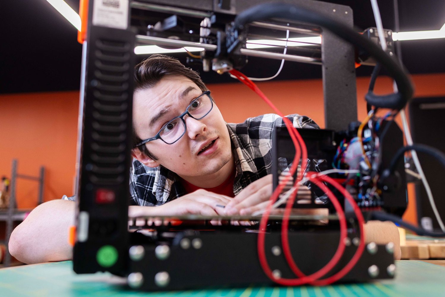 Student sits with arms folded on a table in the Makerspace in front of a 3D printer, which he looks up at with interest and focus