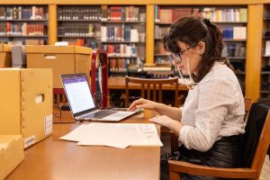Student wearing eyeglasses sits at a table in an archival research room. On the table in front of them sit an open laptop, papers, and boxes of archival materials