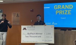 The winner of Biz Pitch is in front of a monitor that reads Grand Prize Xygos holding the big winners check.
