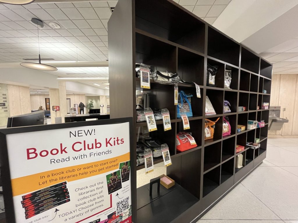A black bookshelf with clear tote bags containing book club kits and buddy read kits. A sign labeled Book Club Kits is at left.