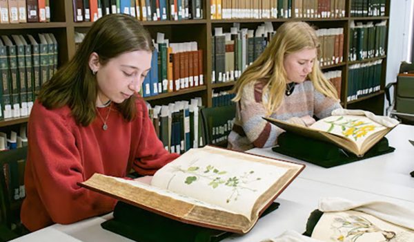 Two female-presenting students sit at a table in a library looking at open books.
