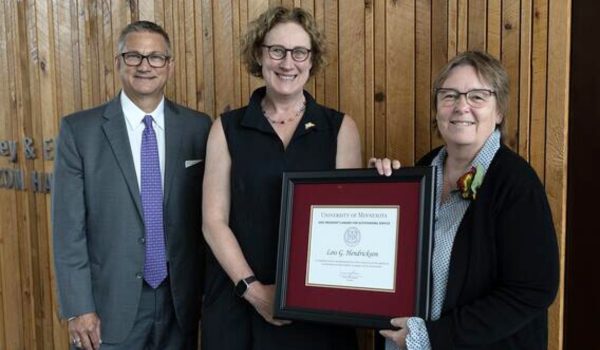 A male-presenting and two female-presenting individuals stand and pose for a photo, presenting a framed certificate.