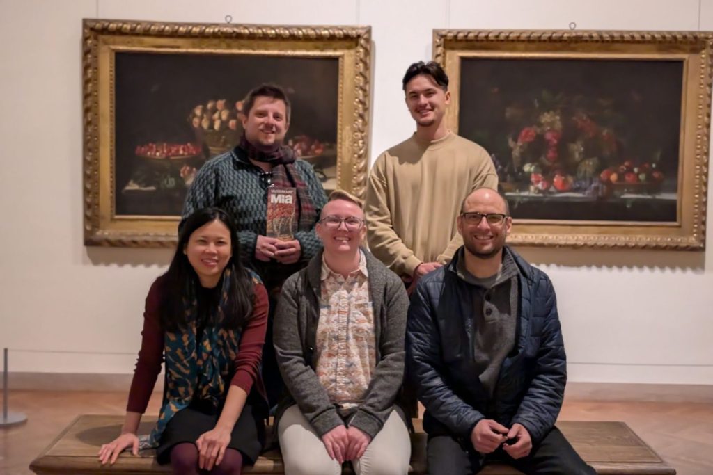 Five individuals posing for a photo in front of two paintings of still life fruit bowls.