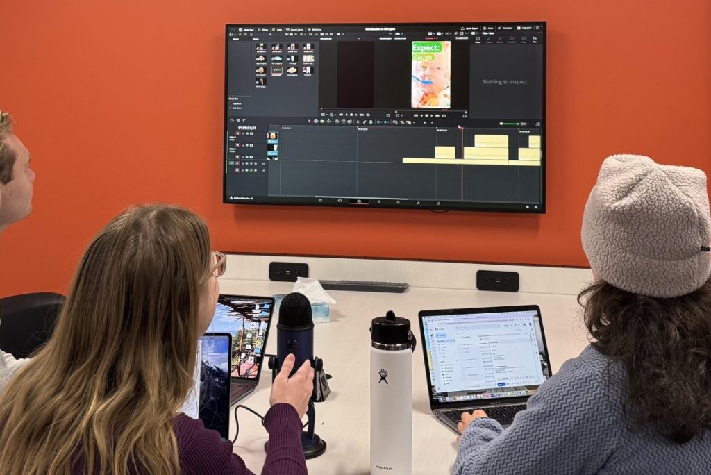 Three students on laptops in front of a screen showing video editing software.