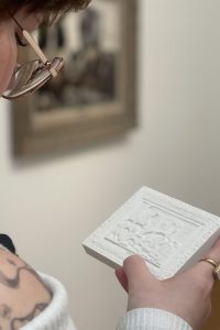 Museum goer holding a 3d printed white tile. The tile pattern is a relief of the artwork the person is standing next to.